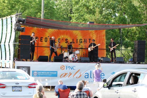 A band on stage with guitars, brass and drums and an orange tie-dye banner that reads "Peoples Light" behind them. 