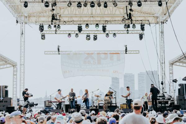 A five-piece band plays on stage on an overcast day. The crowd is in the foreground and a hazy Philadelphia skyline can be seen in the background.