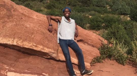 B.J. posing on a hill of orange rock. They are wearing a white top, jeans and a huge smile!